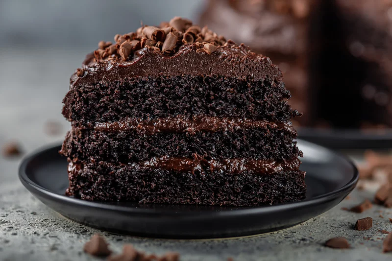 Slice of dark chocolate layer cake on black plate with chocolate curls and frosting, surrounded by scattered chocolate pieces on textured surface