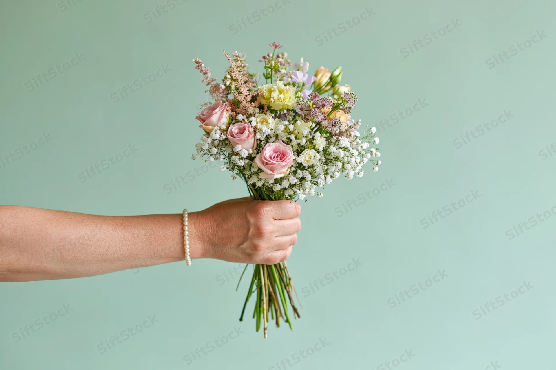 Woman's hand holding a mixed flower bouquet against a mint green background