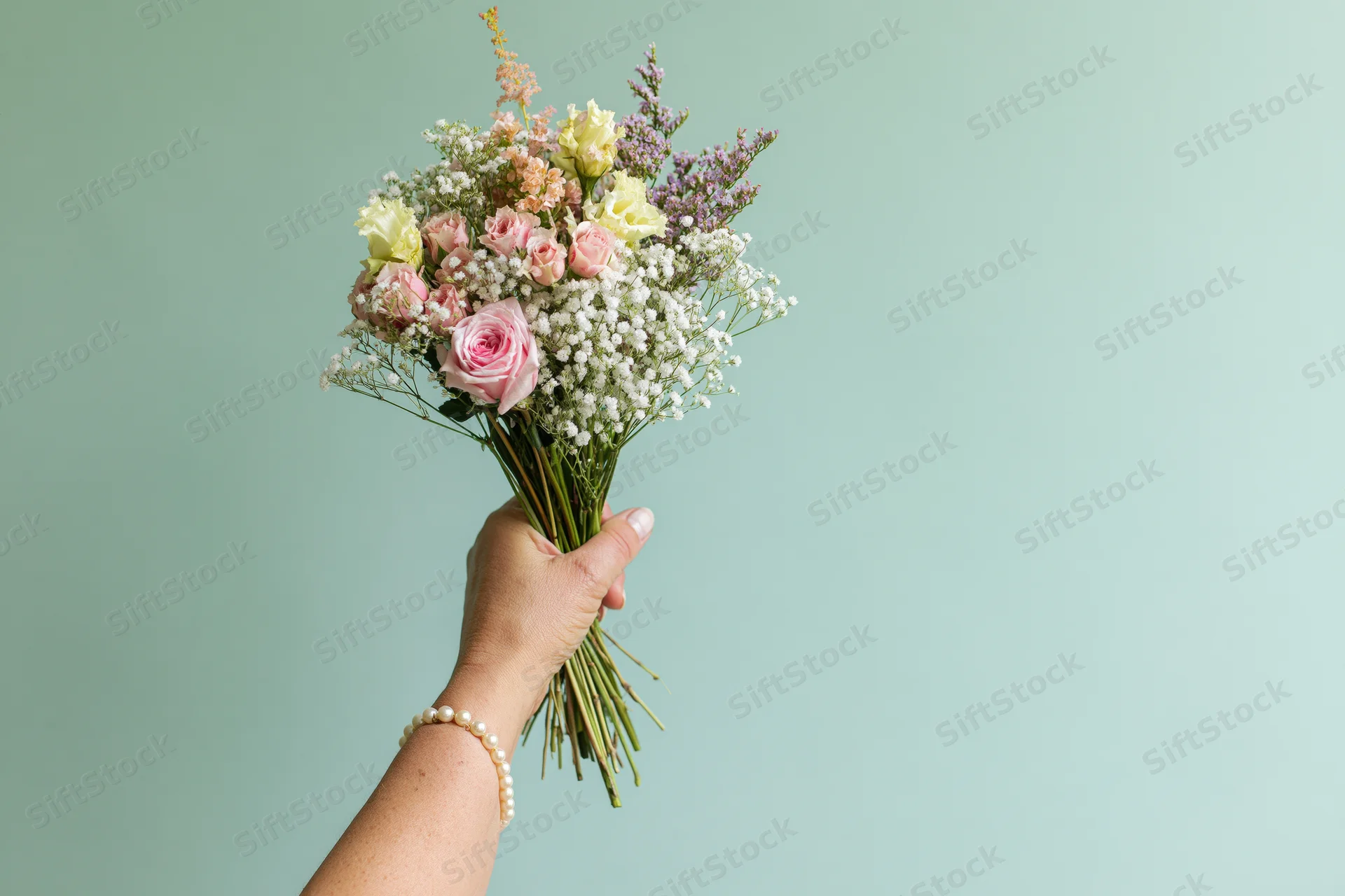 Woman's hand holding a mixed bouquet of pink roses and baby's breath against a mint background