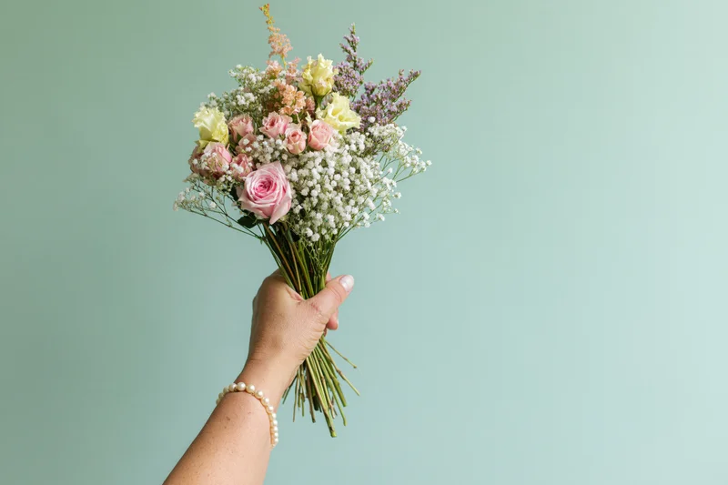 Woman's hand holding a mixed bouquet of pink roses and baby's breath against a mint background