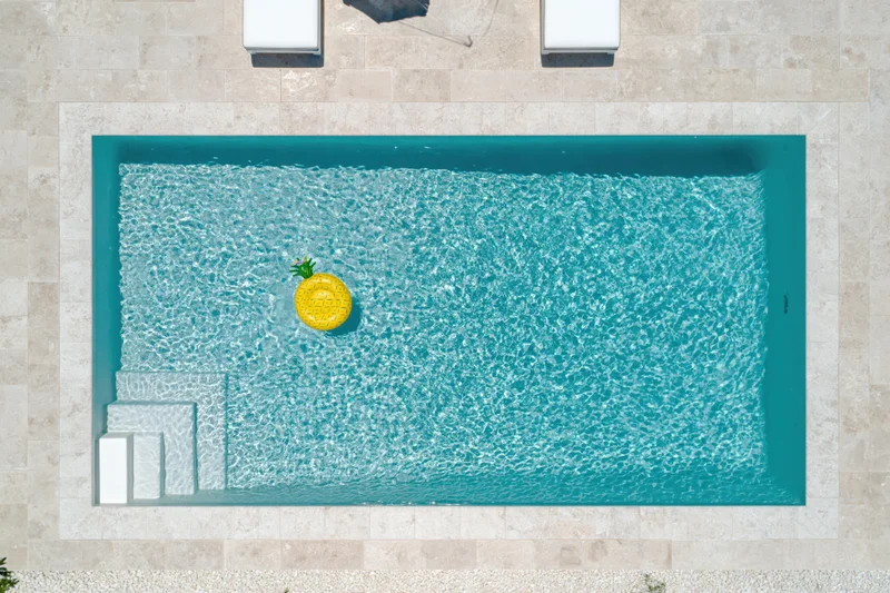 Aerial overhead view of a rectangular outdoor swimming pool with clear turquoise water and a yellow inflatable pineapple floating in it, surrounded by beige stone tiles and white deck chairs