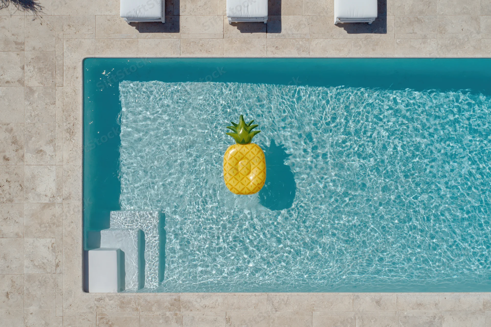 Overhead view of a rectangular swimming pool with a pineapple inflatable floating in clear turquoise water beside white lounge chairs on a tiled deck