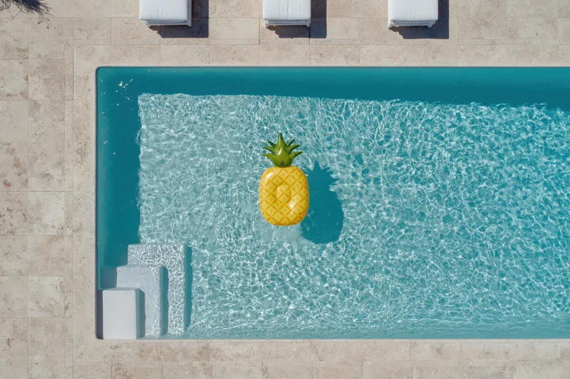 Overhead view of a rectangular swimming pool with a pineapple inflatable floating in clear turquoise water beside white lounge chairs on a tiled deck