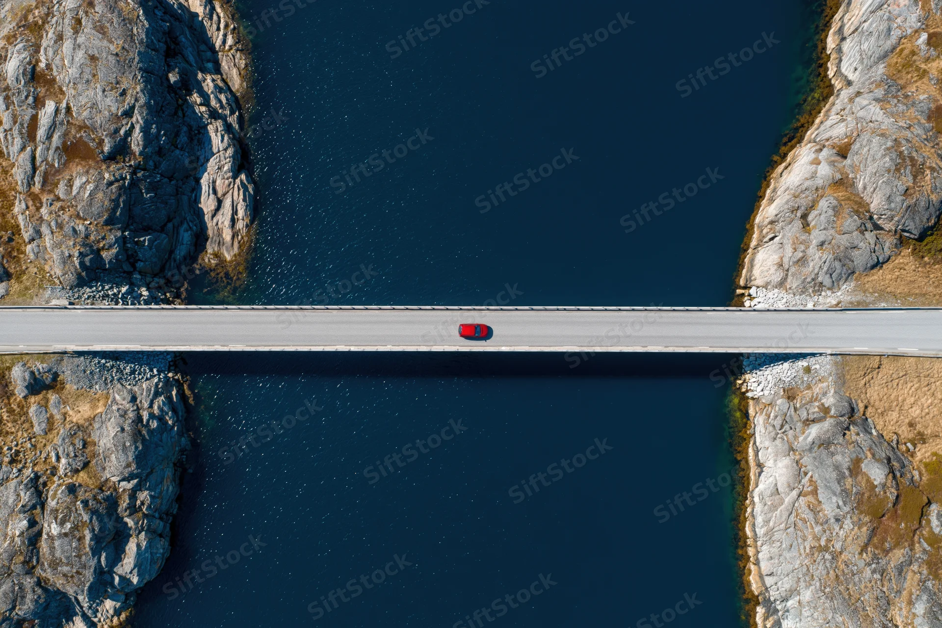 Red car driving across narrow concrete bridge over deep blue water between rocky cliffs, aerial top-down view