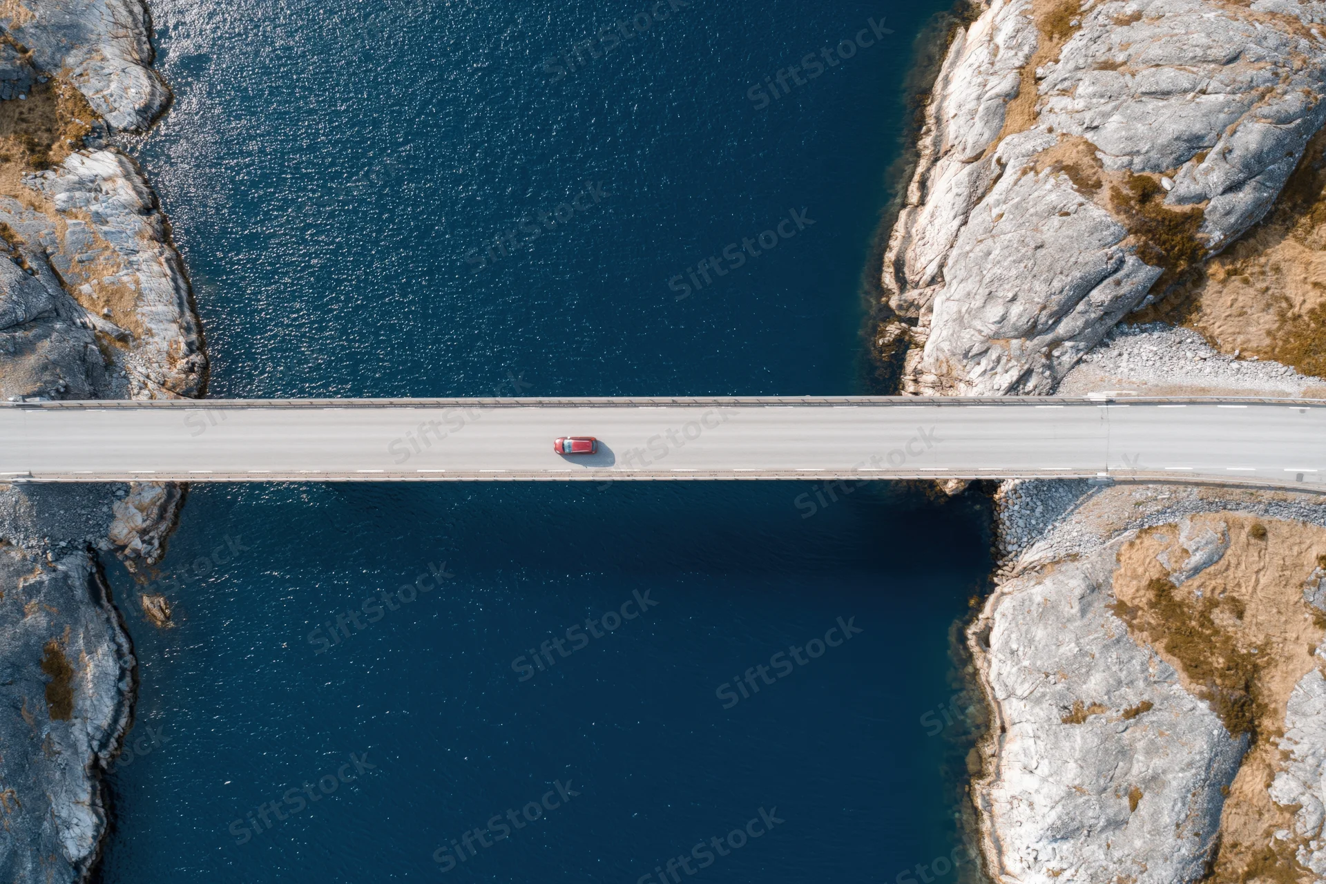 Aerial top-down view of a red car driving across a narrow concrete bridge spanning deep blue water between rocky cliffs