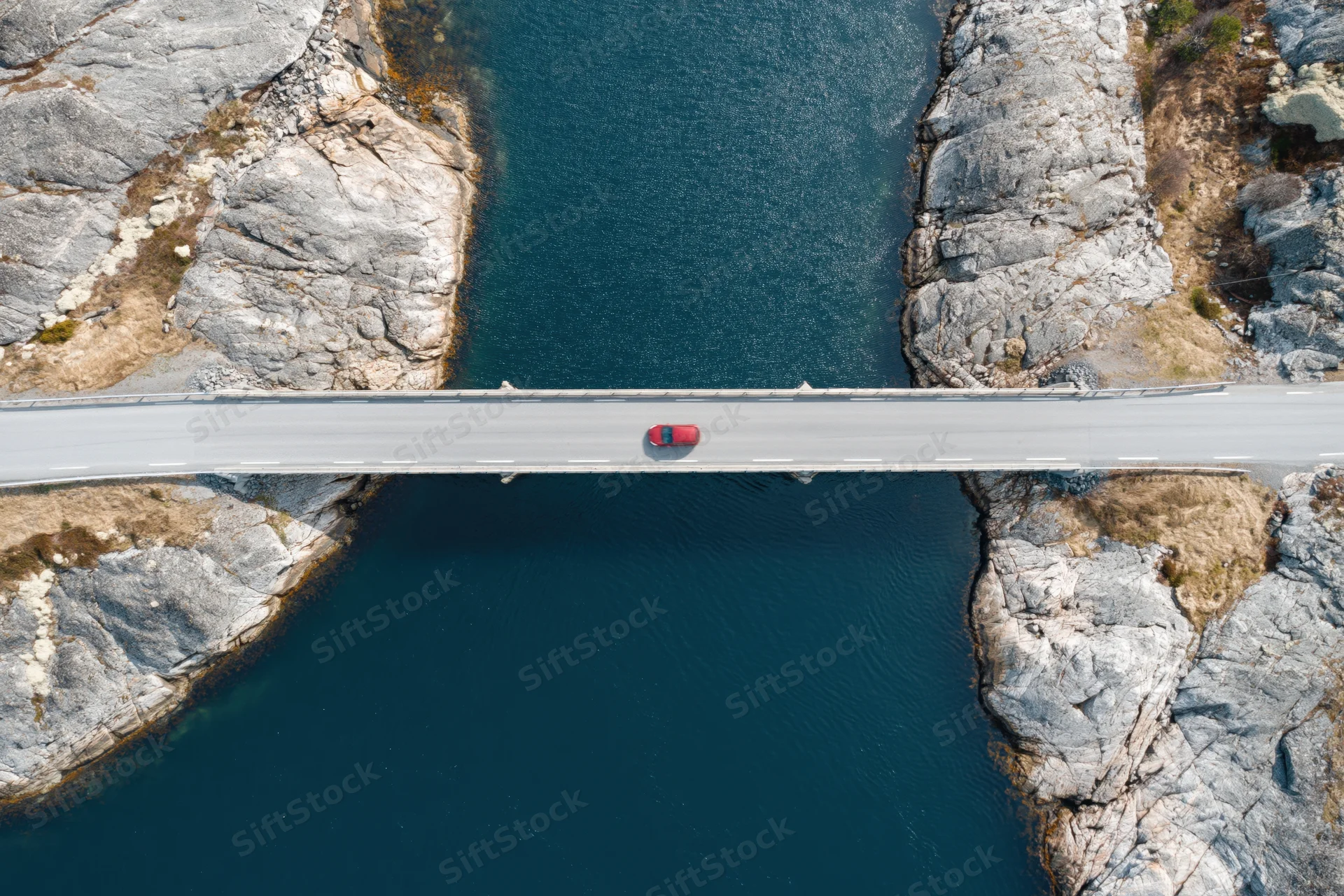 Red car driving across narrow concrete bridge over deep blue water between rocky cliffs