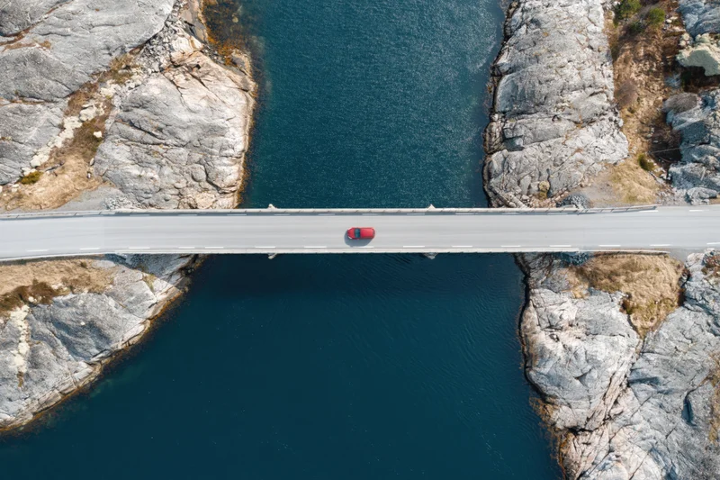 Red car driving across narrow concrete bridge over deep blue water between rocky cliffs
