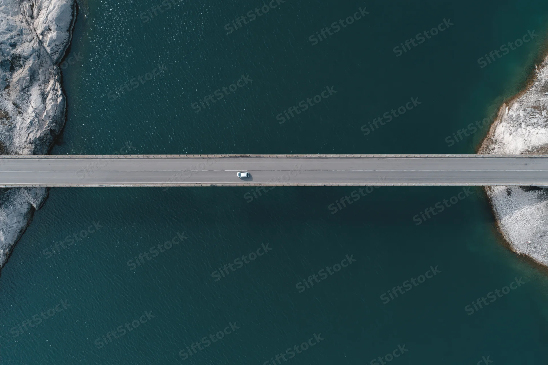 Aerial top-down view of a car driving across a narrow bridge over a deep blue river flanked by snow-covered rocky shores