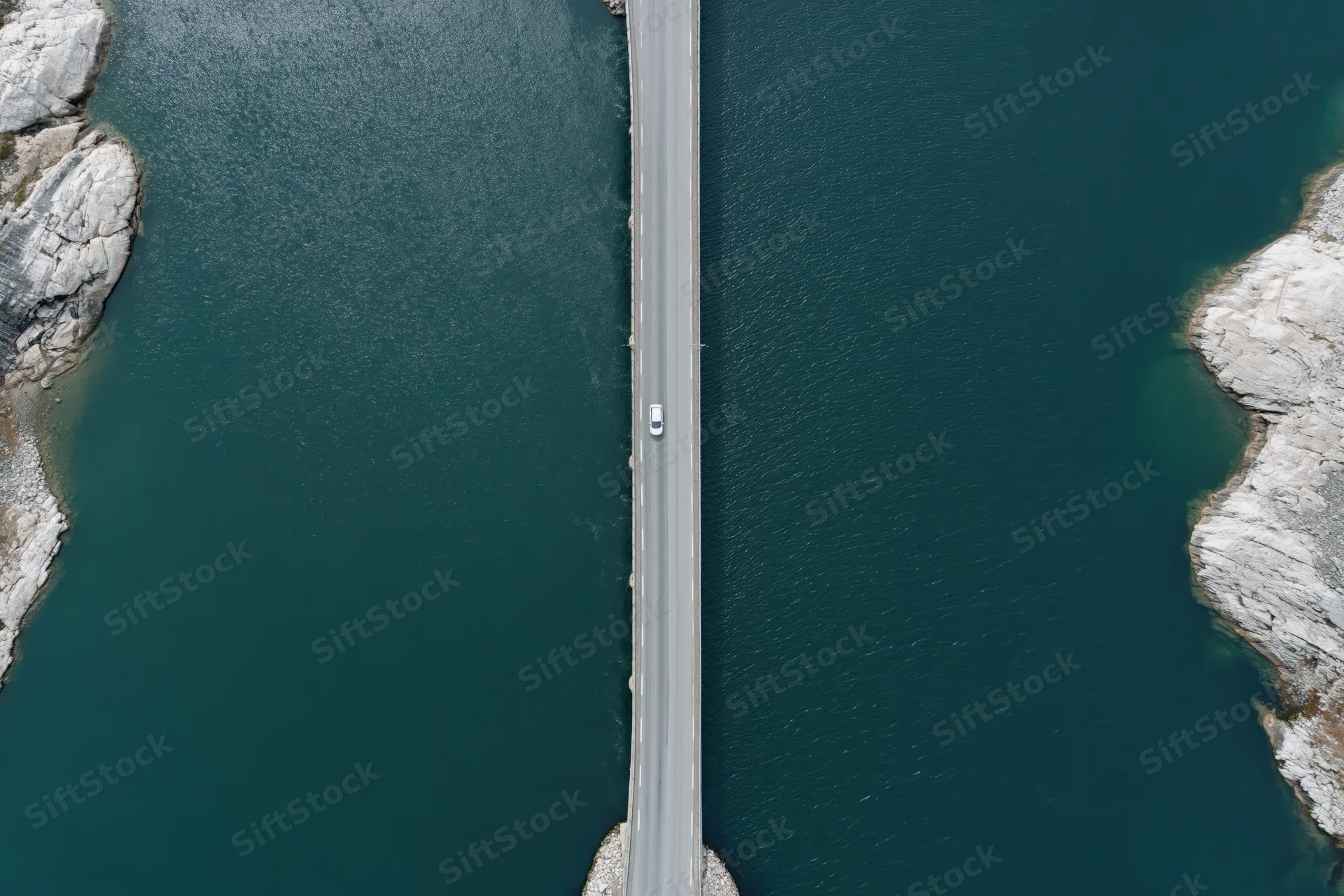 Aerial top-down view of a narrow road bridge over dark teal water with a white car driving across, flanked by rocky cliffs