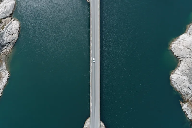Aerial top-down view of a narrow road bridge over dark teal water with a white car driving across, flanked by rocky cliffs