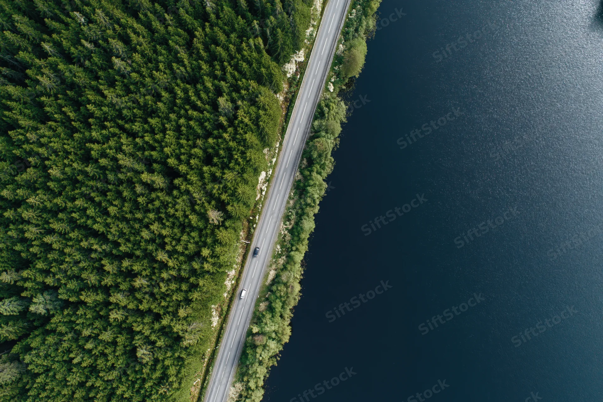 Aerial top-down view of a narrow two-lane road running between dense green forest and dark blue lake with cars driving along it