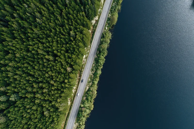 Aerial top-down view of a narrow two-lane road running between dense green forest and dark blue lake with cars driving along it