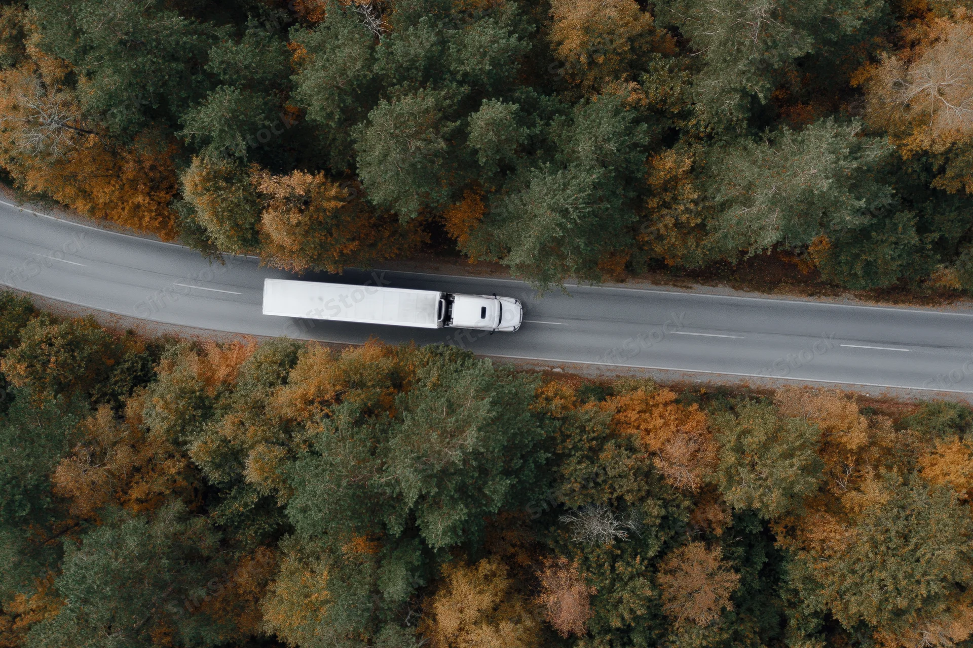 White semi-truck driving on a highway surrounded by autumn forest from aerial top-down view