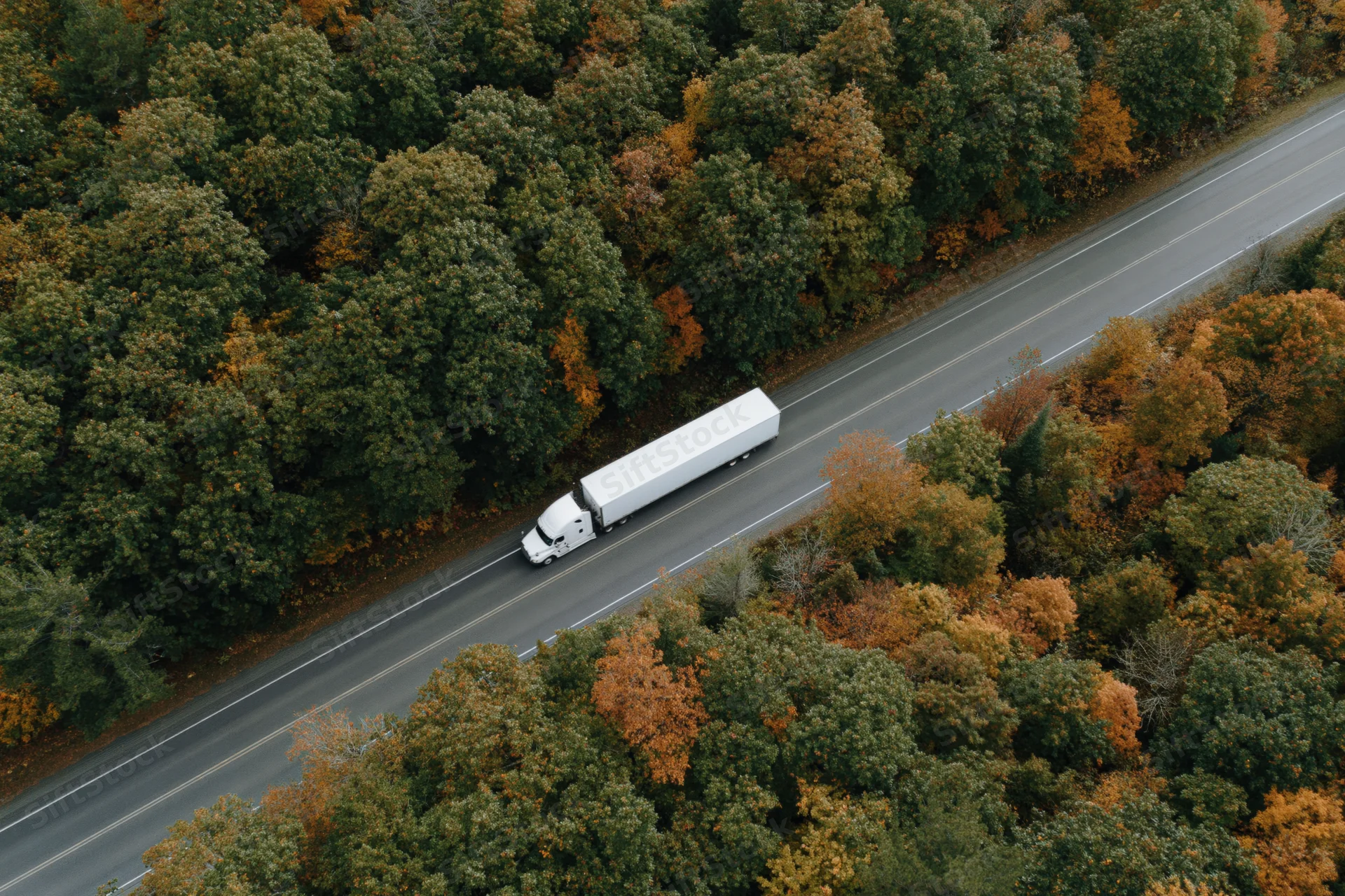White semi-truck driving on highway surrounded by autumn forest from aerial view