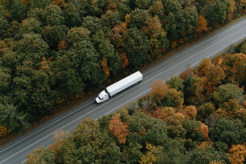 White semi-truck driving on highway surrounded by autumn forest from aerial view