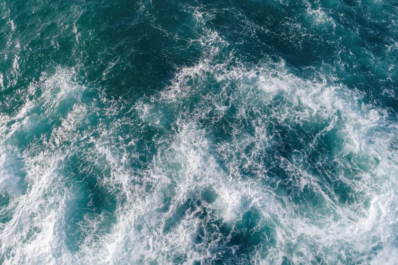Aerial top-down view of open ocean surface with scattered white foam and swirling waves