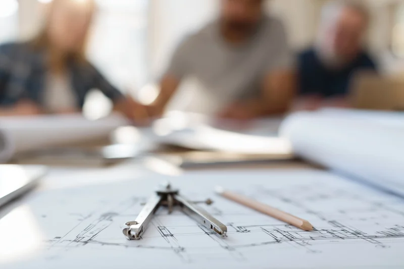 Drafting compass and pencil on blueprint with team in background at office meeting