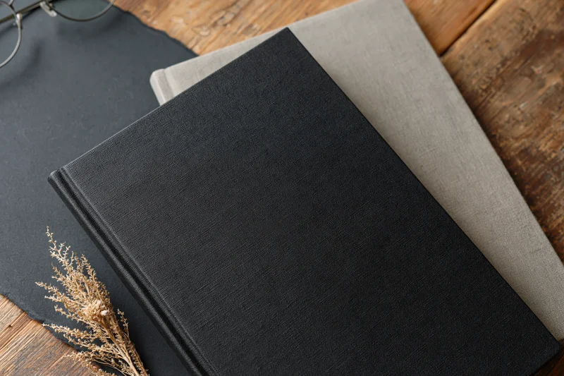 Black hardcover notebook with textured matte surface resting on wooden table beside dried grass and eyeglasses