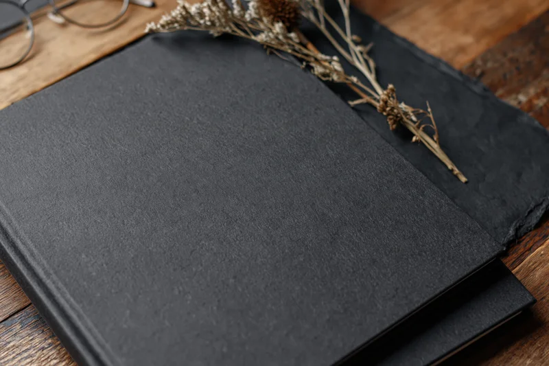 Black hardcover notebook with textured matte surface resting on wooden table with dried flowers and eyeglasses nearby
