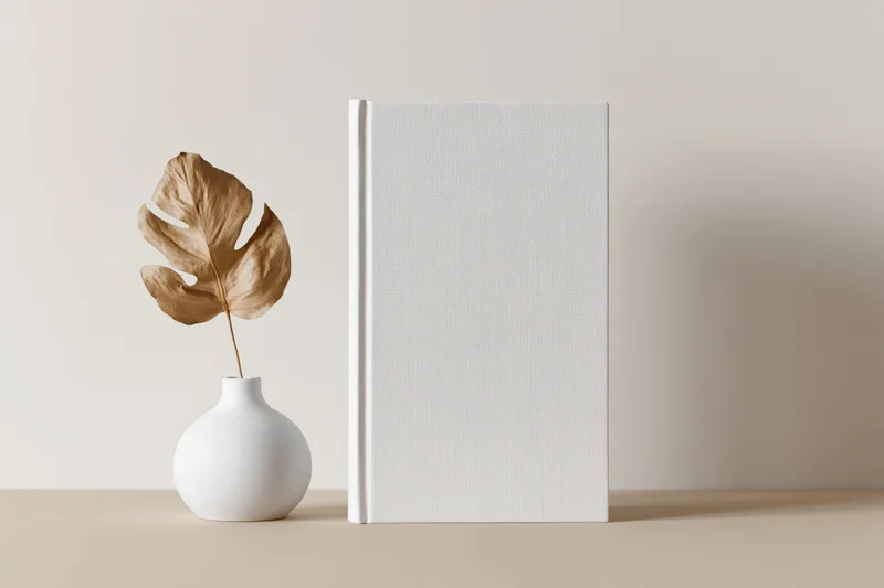 Blank white hardcover book standing upright next to a small white vase with a dried monstera leaf on a beige surface