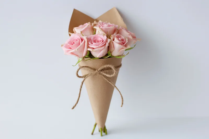 Bouquet of soft pink roses wrapped in kraft paper cone tied with twine, standing on white surface