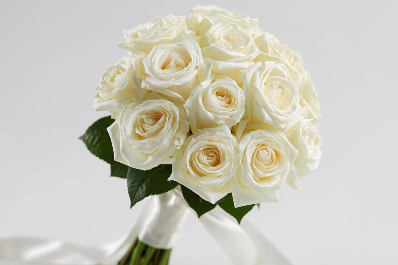 Bridal bouquet of tightly clustered white roses with green leaves and white ribbon, held upright against white background