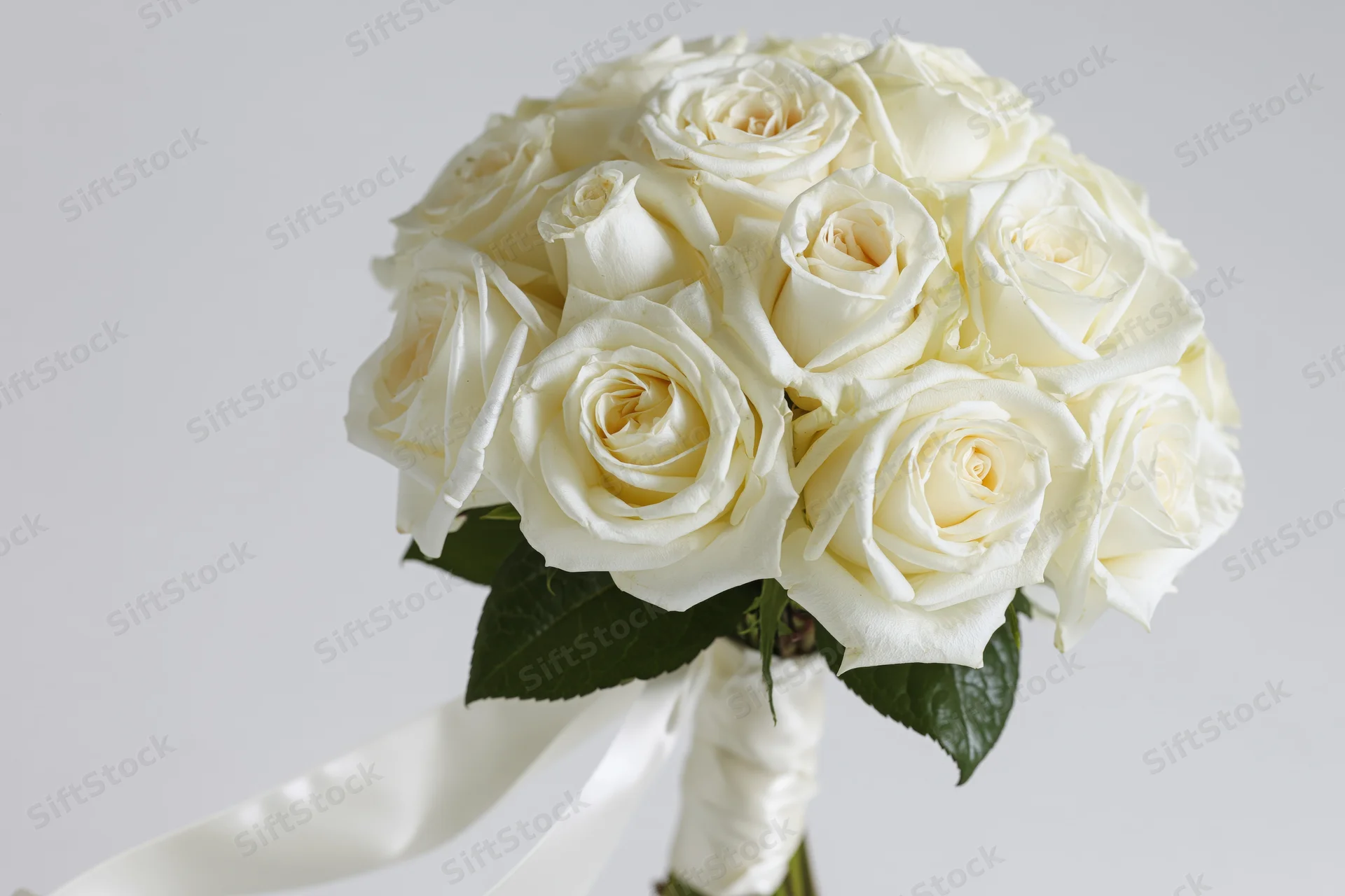 Bridal bouquet of tightly clustered white roses with green leaves and satin ribbon, held upright against white background