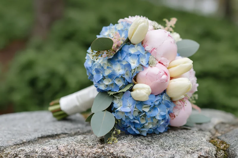 Bridal bouquet with blue hydrangeas, pink peonies and cream tulips resting on a rough stone surface outdoors