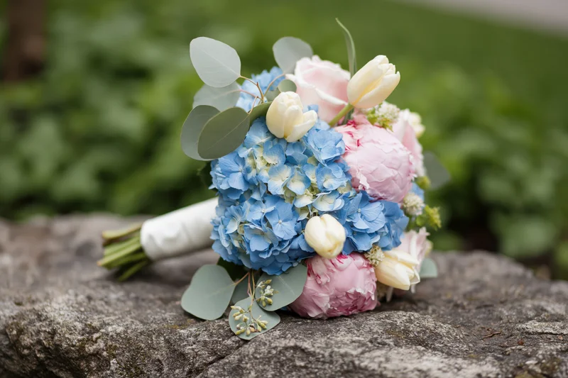 Bridal bouquet with blue hydrangeas, pink peonies and white tulips resting on a rough stone outdoors