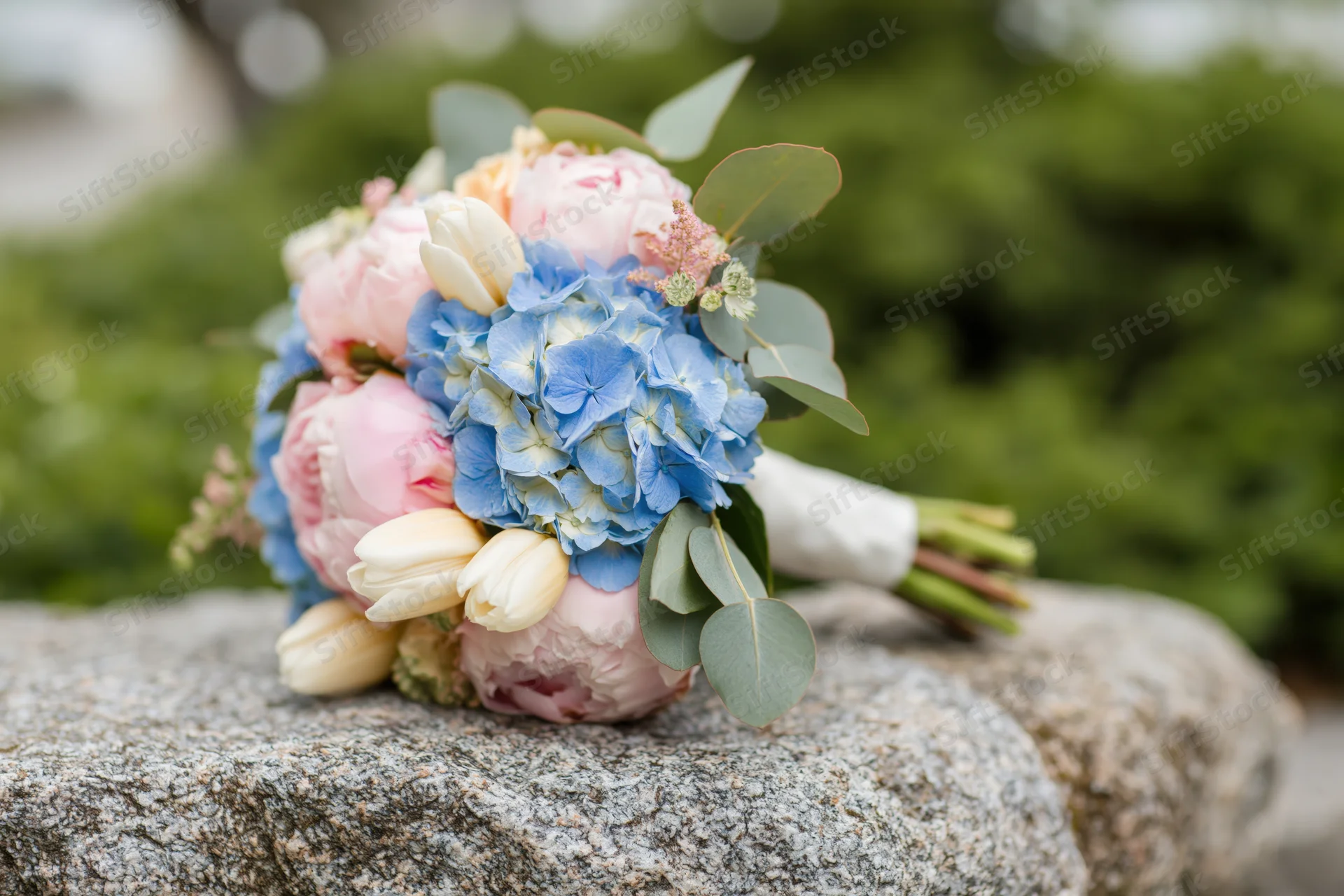Bridal bouquet with pink peonies, blue hydrangeas and white tulips resting on a rough stone outdoors