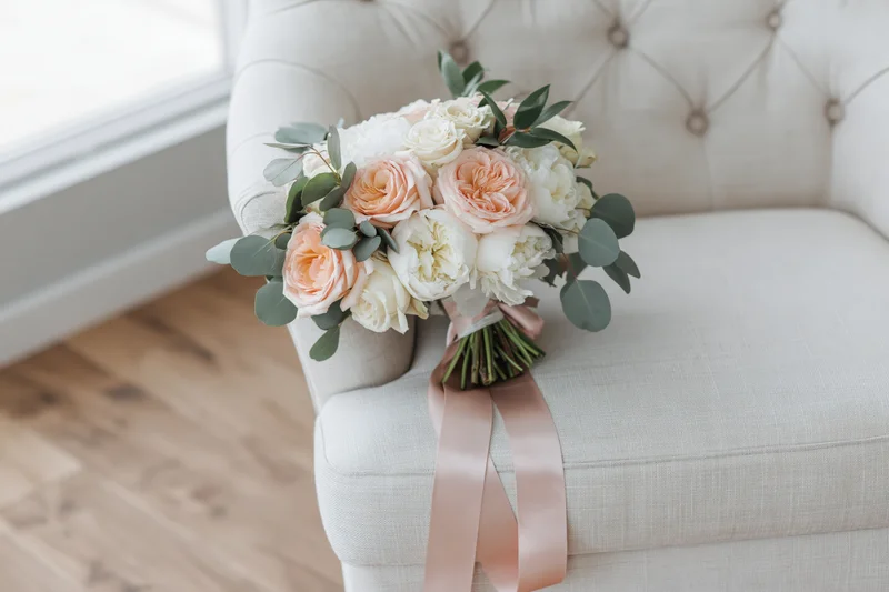 Bridal bouquet with peach roses and white peonies resting on tufted cream linen armchair