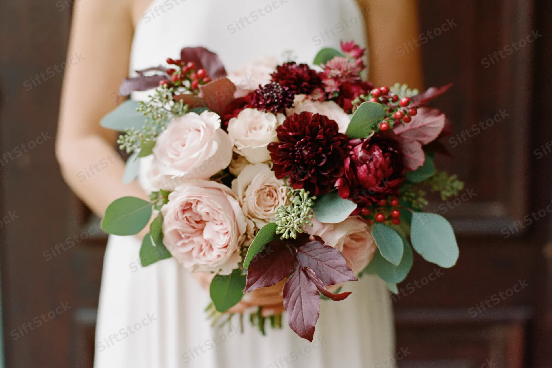 Bride holding a lush wedding bouquet of blush pink garden roses and burgundy flowers in front of a wooden door