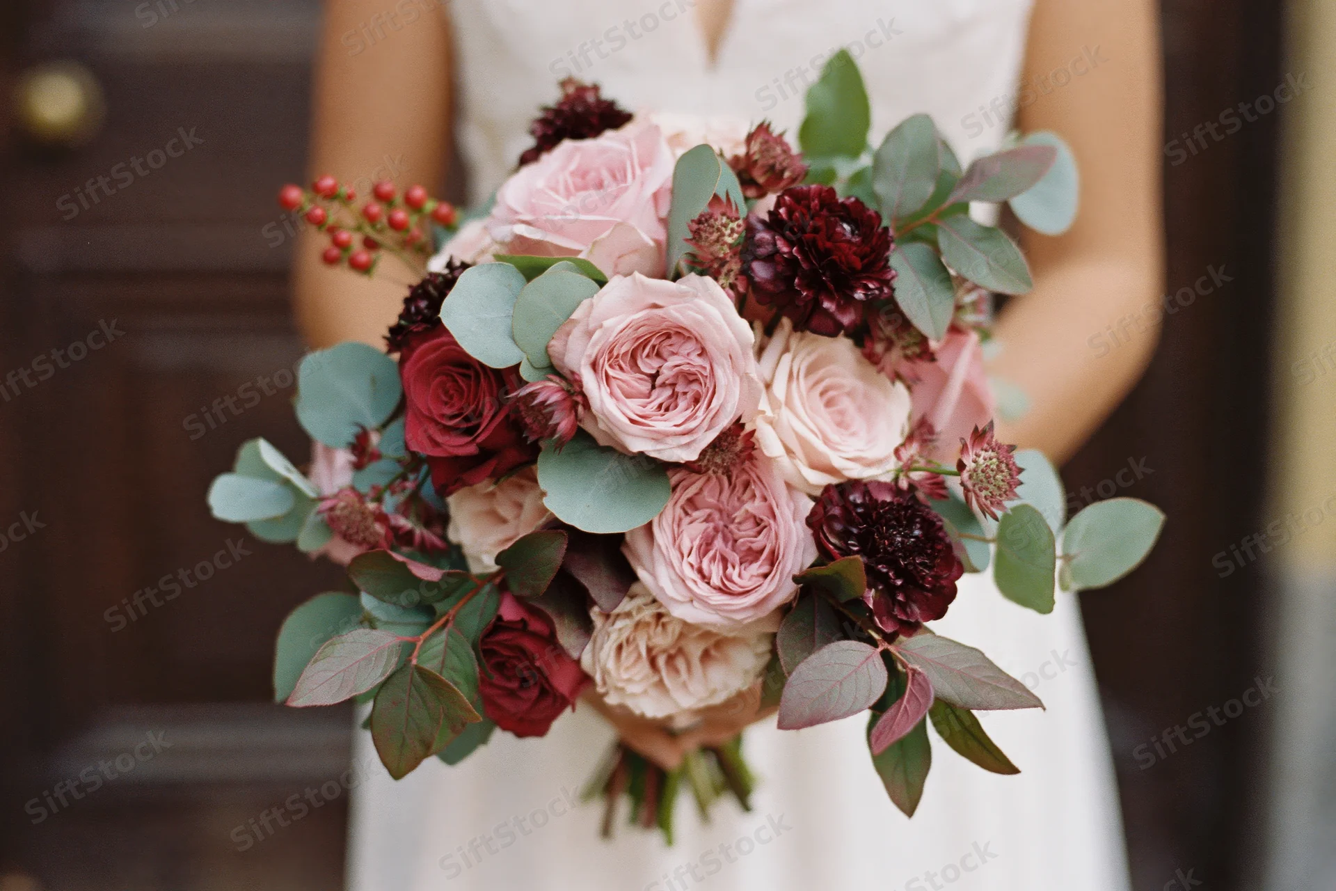 Bride holding a lush wedding bouquet of blush pink garden roses and burgundy flowers with green foliage in front of a wooden door