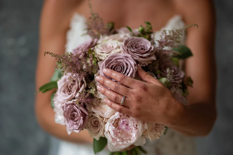 Bride holding a dusty mauve and blush rose bouquet with peonies and greenery, hand resting on flowers showing wedding ring