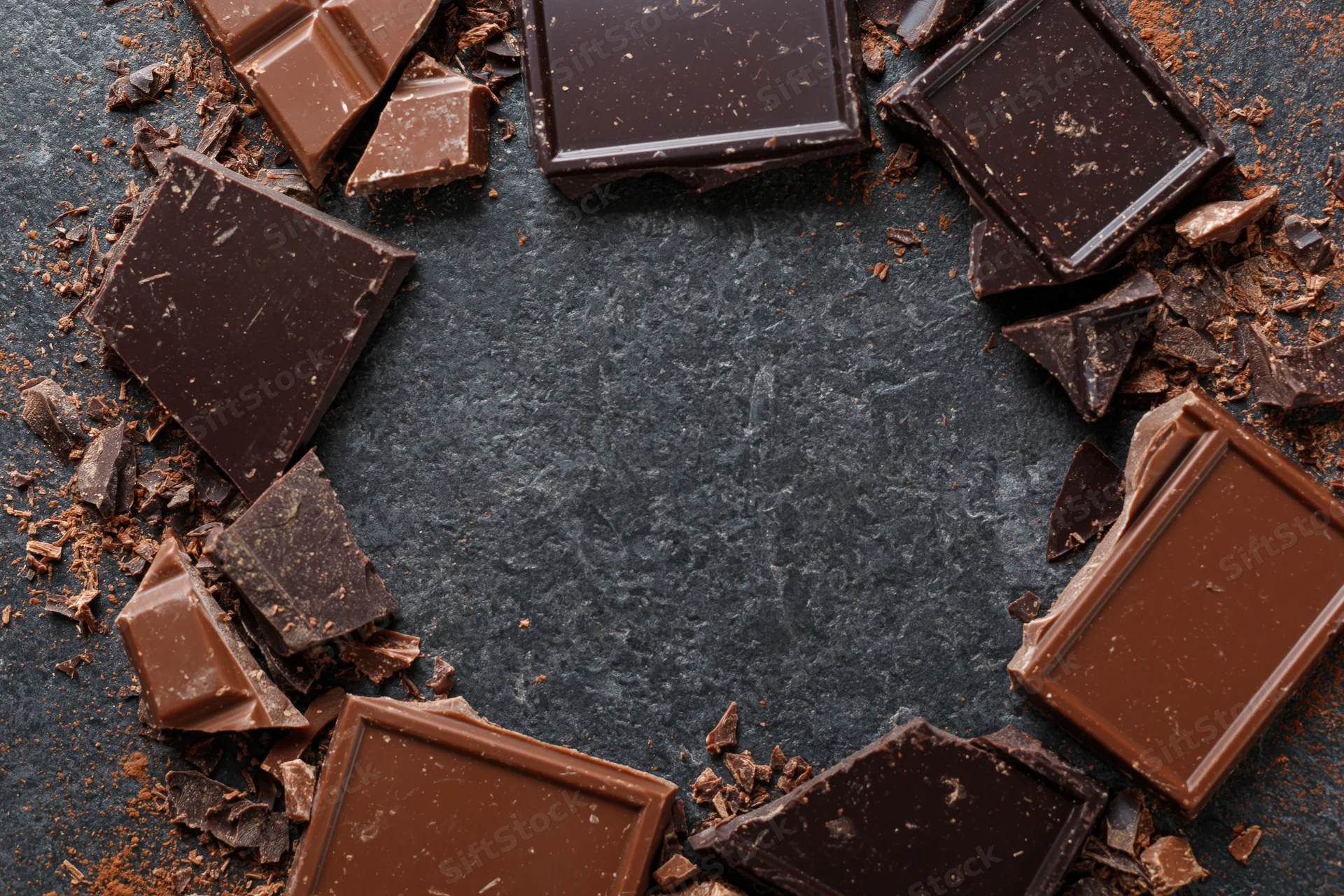 Broken dark and milk chocolate pieces arranged in a loose circular frame on a dark slate surface with central copyspace