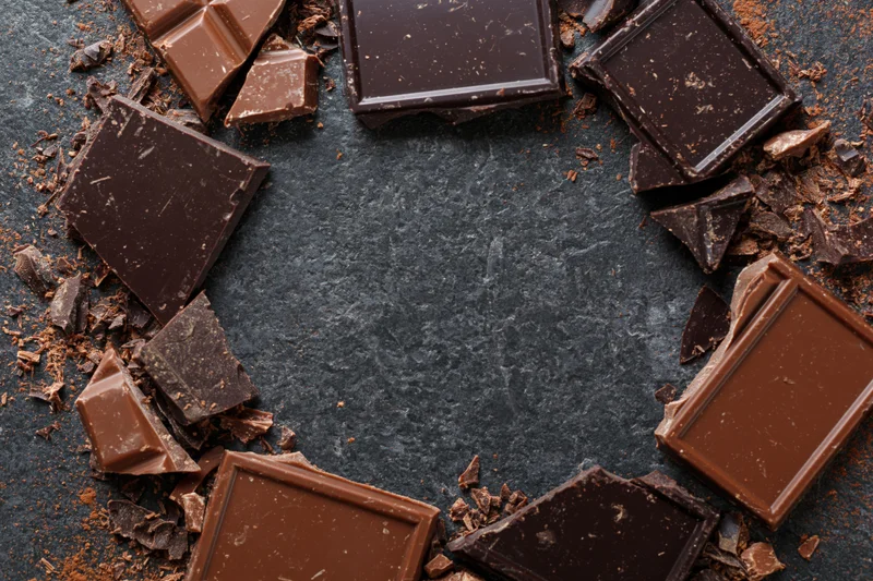Broken dark and milk chocolate pieces arranged in a loose circular frame on a dark slate surface with central copyspace