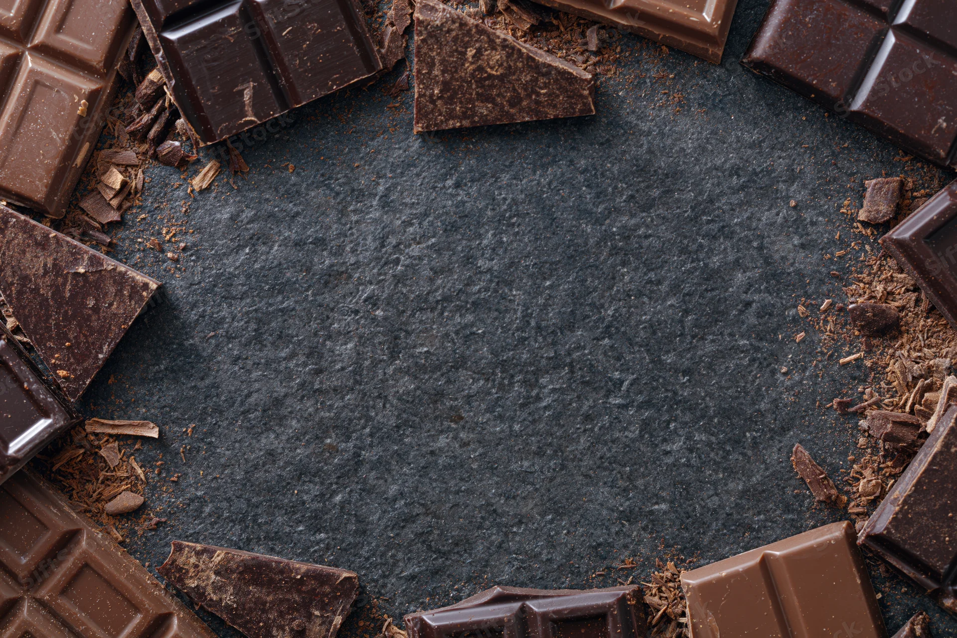 Broken dark and milk chocolate pieces arranged in a loose circular frame on a dark slate surface with central copyspace