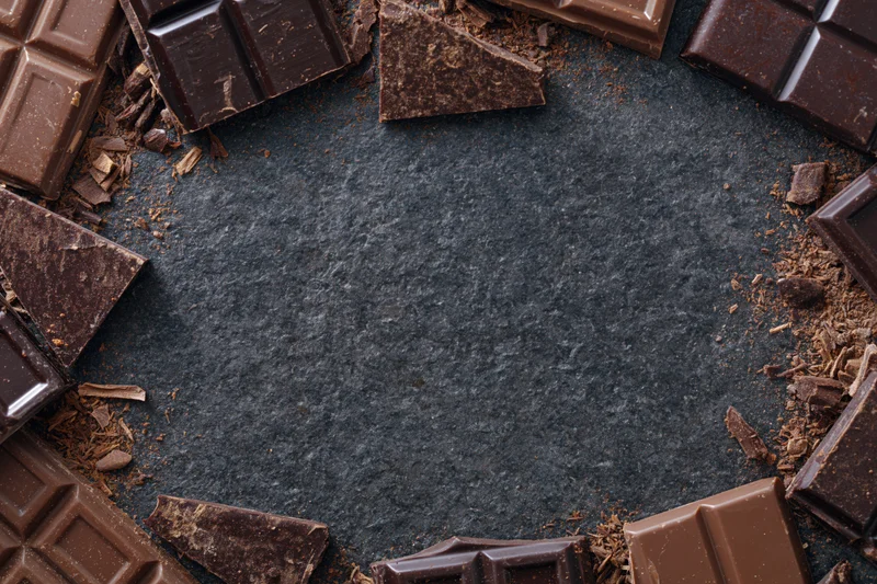Broken dark and milk chocolate pieces arranged in a loose circular frame on a dark slate surface with central copyspace