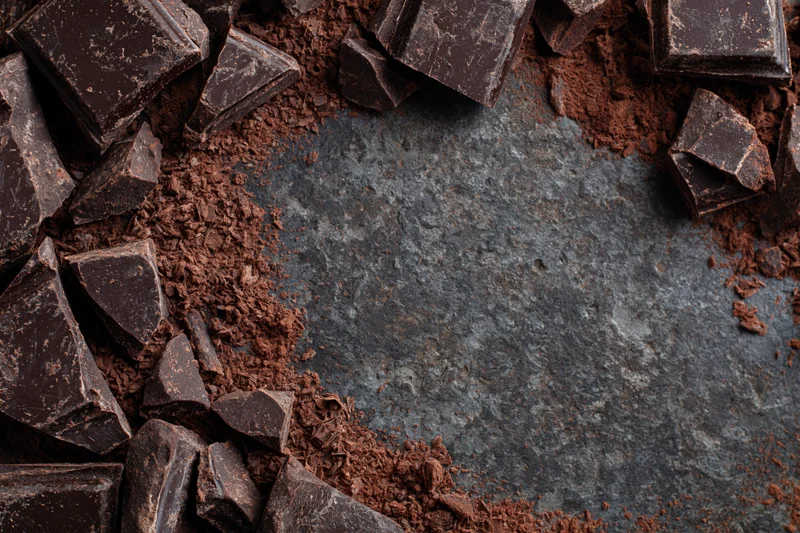 Broken dark chocolate chunks and cocoa shavings arranged in a circular border on a slate surface with central copyspace