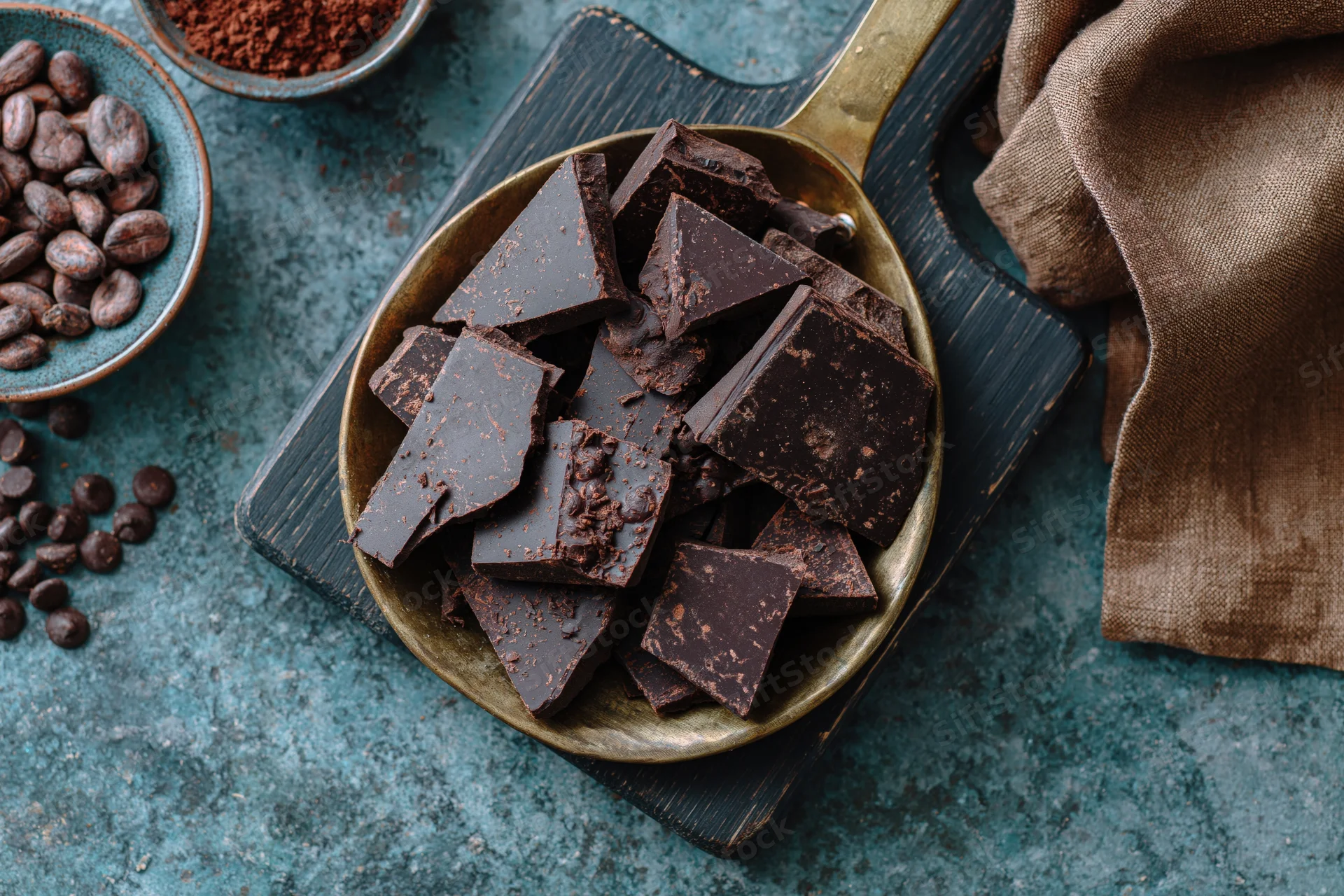 Broken dark chocolate chunks in a vintage brass pan on a wooden cutting board with cocoa beans and powder nearby