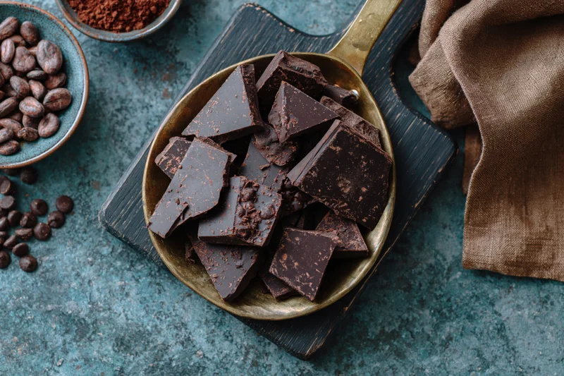 Broken dark chocolate chunks in a vintage brass pan on a wooden cutting board with cocoa beans and powder nearby