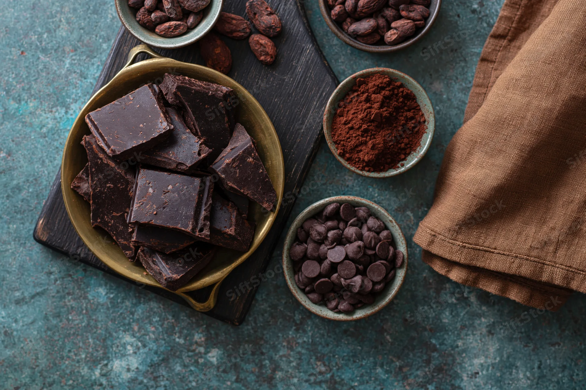 Broken dark chocolate chunks in a vintage brass pan with cocoa beans, powder, and chips arranged on a wooden board and teal surface