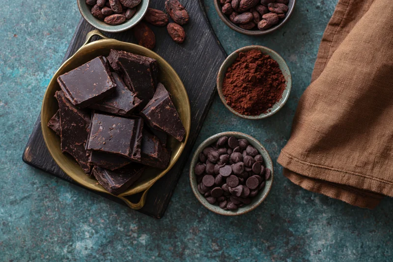 Broken dark chocolate chunks in a vintage brass pan with cocoa beans, powder, and chips arranged on a wooden board and teal surface