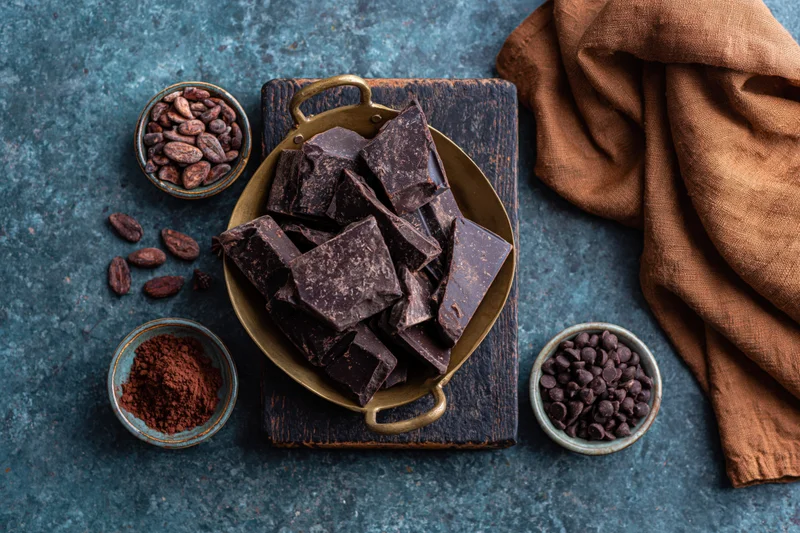 Broken dark chocolate chunks in a vintage brass pan on a wooden board with cocoa beans, powder, and chips around