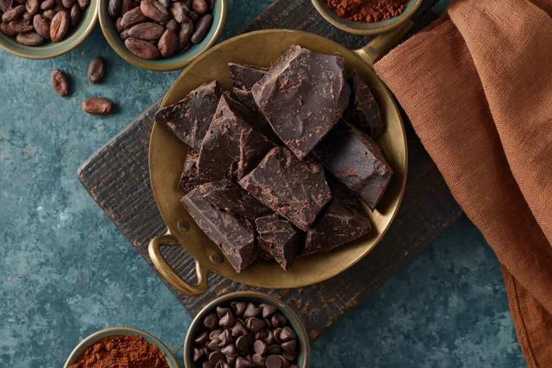 Broken dark chocolate chunks piled in a vintage brass pan on a wooden cutting board with cocoa beans and powder in bowls around it