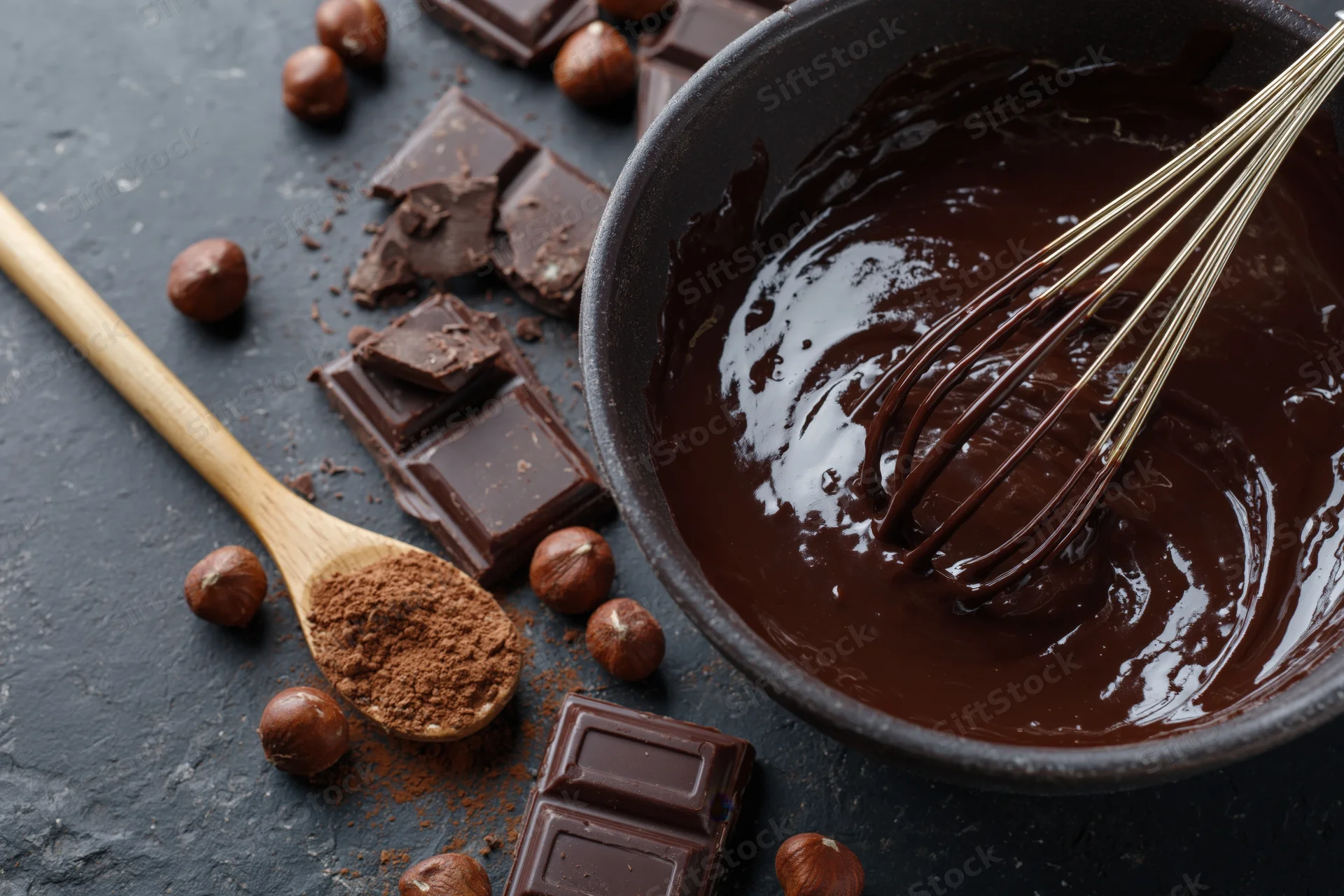 Melted chocolate in a dark ceramic bowl with whisk, surrounded by chocolate pieces, hazelnuts, and cocoa powder on a dark surface