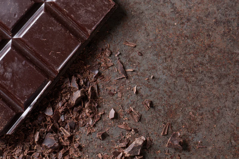 Dark chocolate bar broken into pieces on a textured metal surface with scattered shards and crumbs
