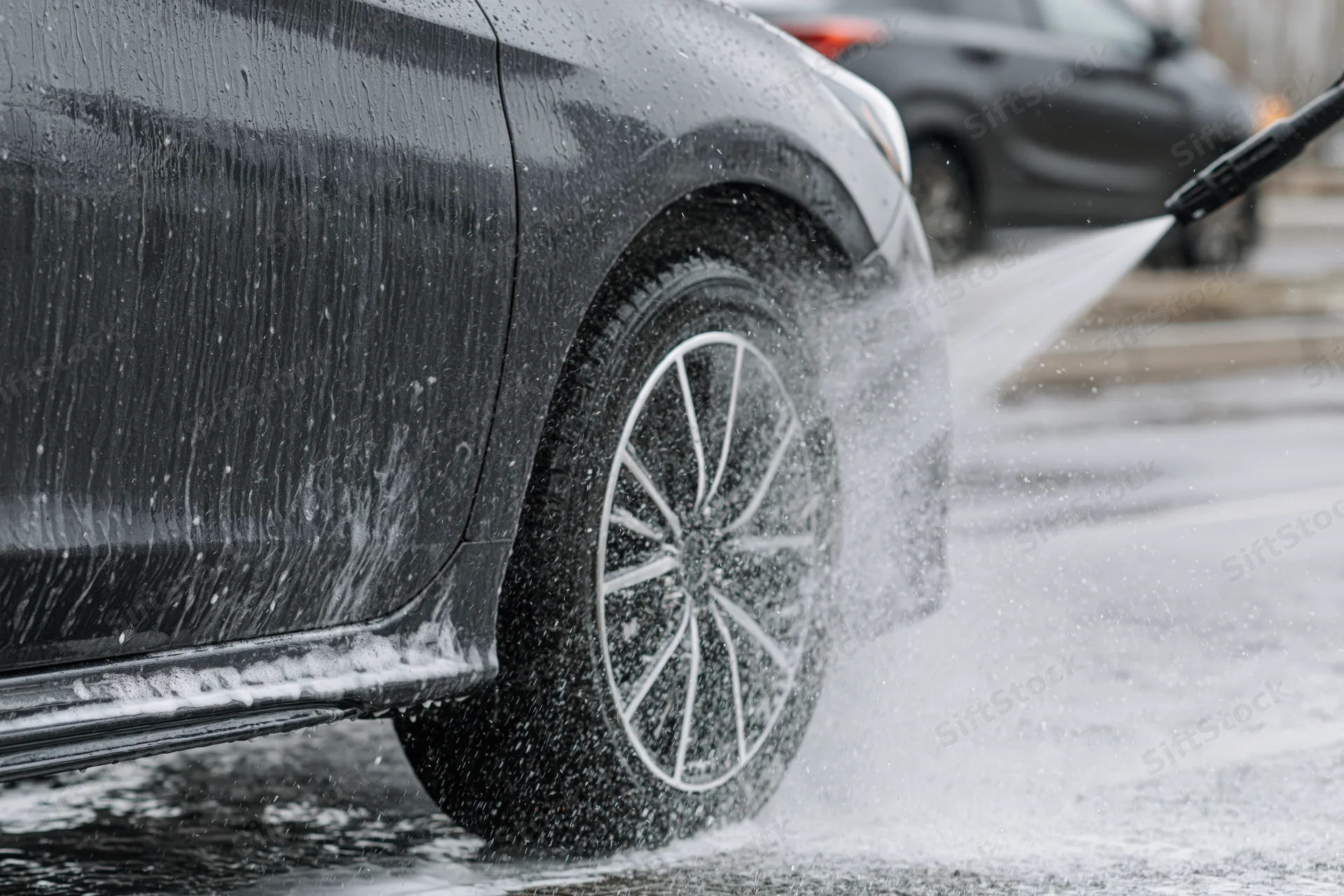 Dark grey sedan being sprayed with pressure washer wand on wet pavement