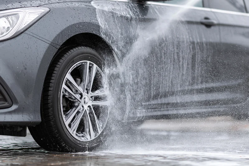 Dark grey sedan being sprayed with pressure washer water on wet pavement