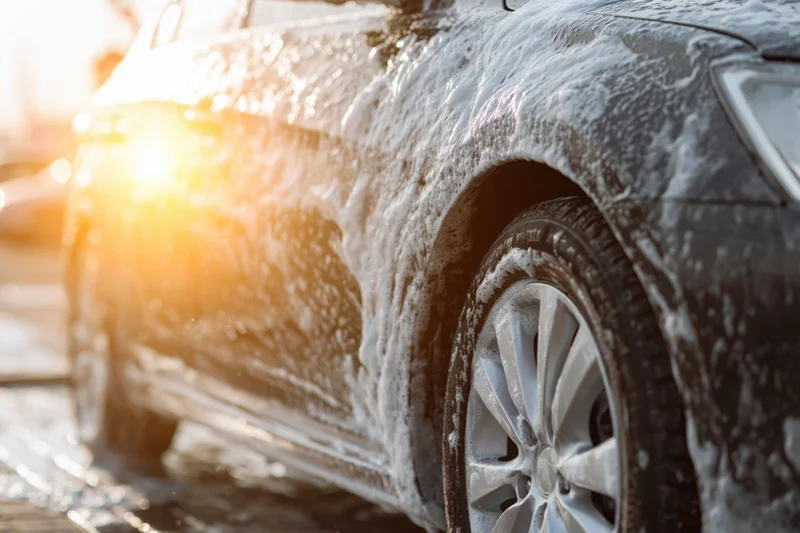 Dark sedan covered in white soapy foam being washed outdoors in sunlight